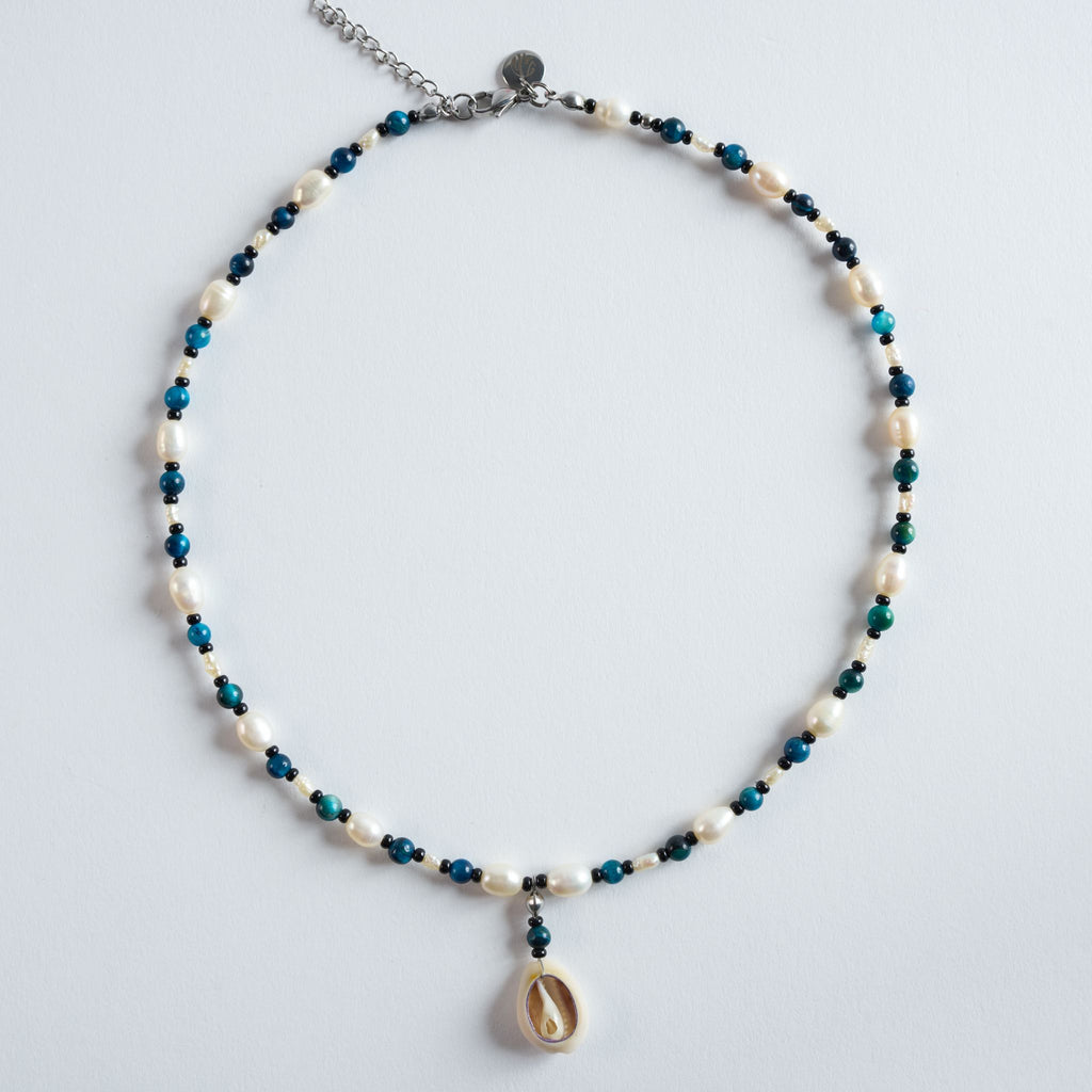 Die Halskette „Deep Ocean“ kombiniert blau gefärbtes Tigerauge mit weichen Süßwasserperlen und einem Muschelanhänger.