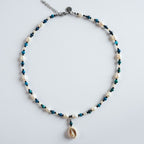Die Halskette „Deep Ocean“ kombiniert blau gefärbtes Tigerauge mit weichen Süßwasserperlen und einem Muschelanhänger.
