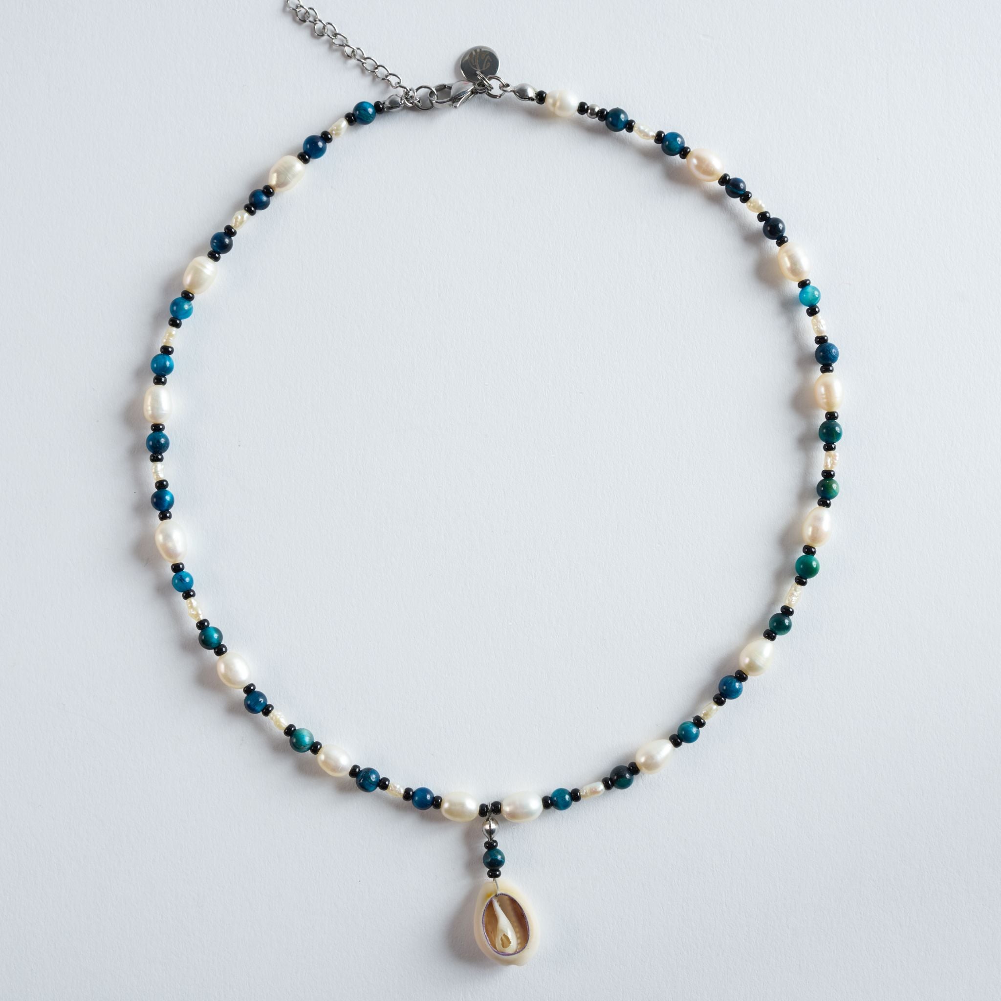 Die Halskette „Deep Ocean“ kombiniert blau gefärbtes Tigerauge mit weichen Süßwasserperlen und einem Muschelanhänger.